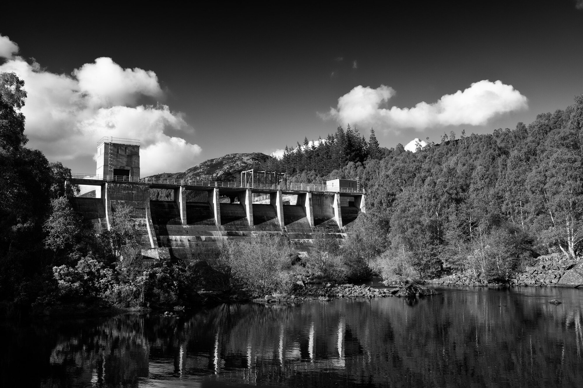 A dam and its towers above still water, bright clouds overhead