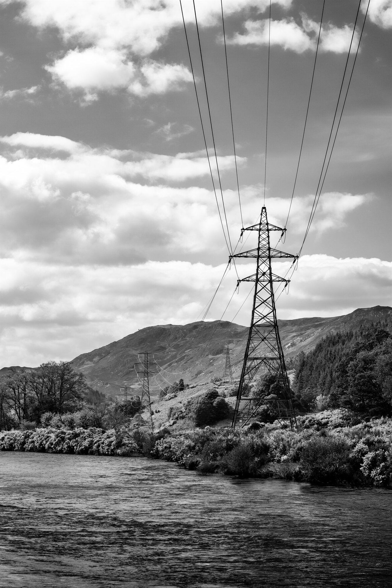 A pylon above a river bank, wires running up into the clouds