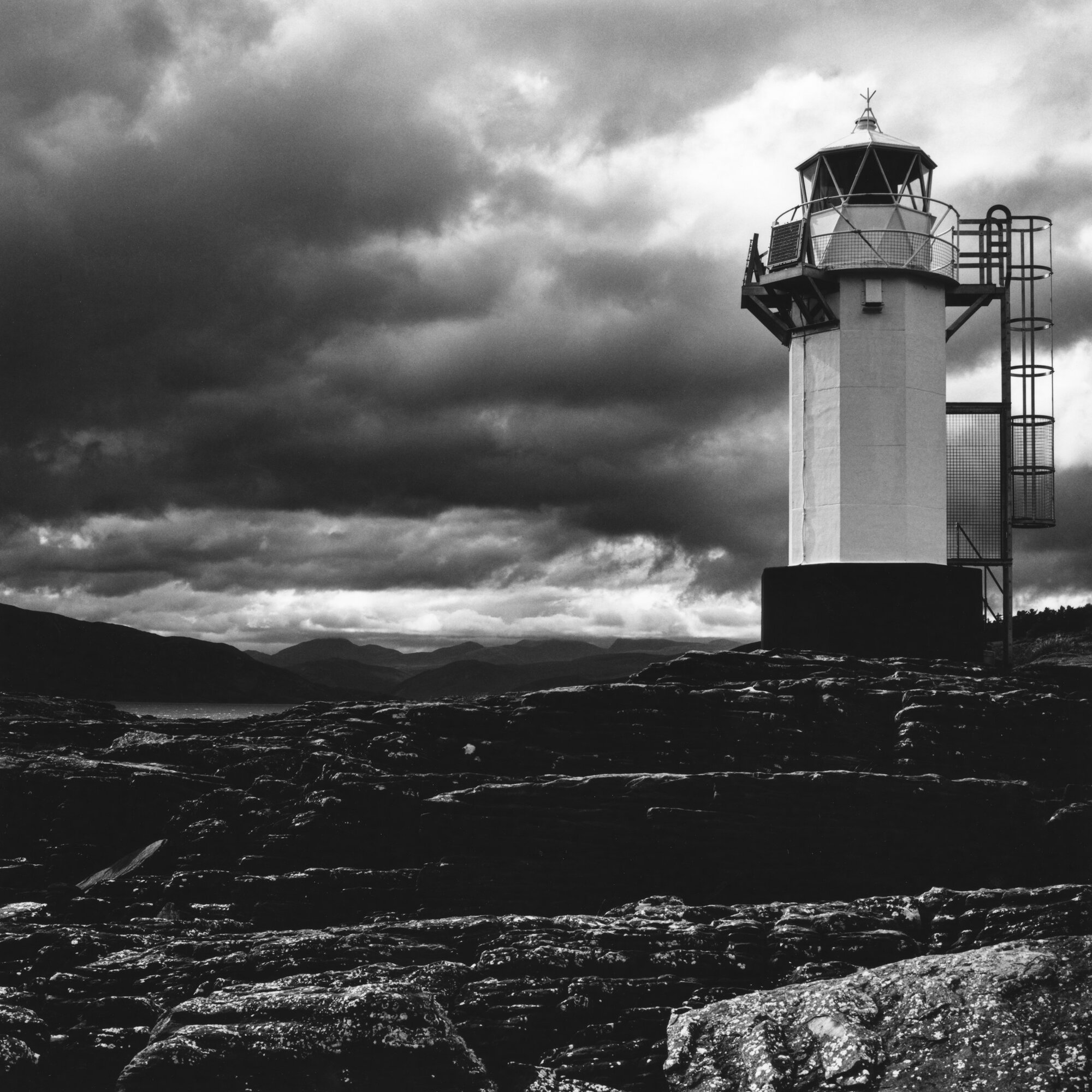 Ullapool lighthouse on the rocks beneath a storm sky, mountains across the loch