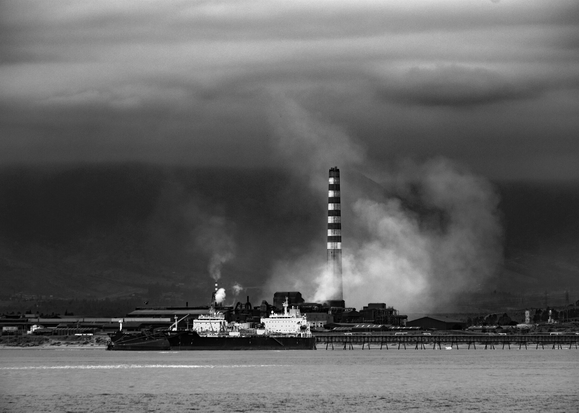 A tall chimney and plume of smoke over the sea under a stormy sky