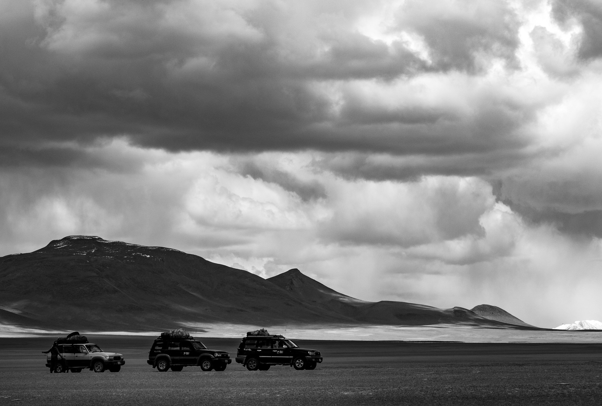 Three loaded jeeps parked on a high plain beneath dark storm clouds and mountains, in black and white